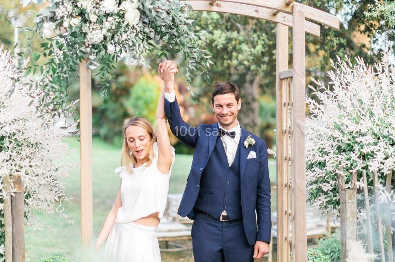 Un couple souriant sous une arche décorée de feuillage, célébrant un moment festif en extérieur.