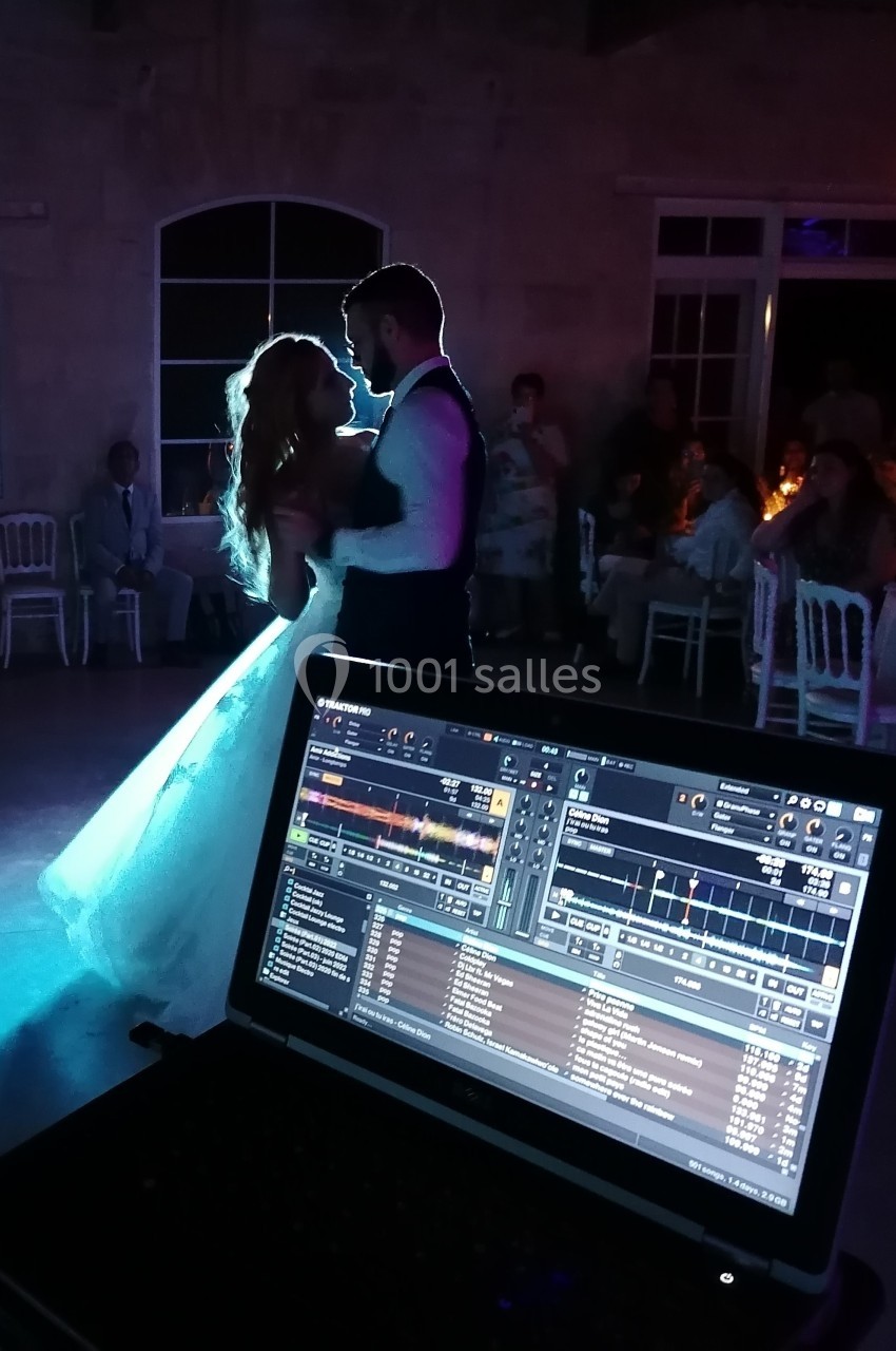 Un couple danse lors d'une réception de mariage, éclairé par une lumière tamisée, avec un ordinateur de DJ au premier plan.