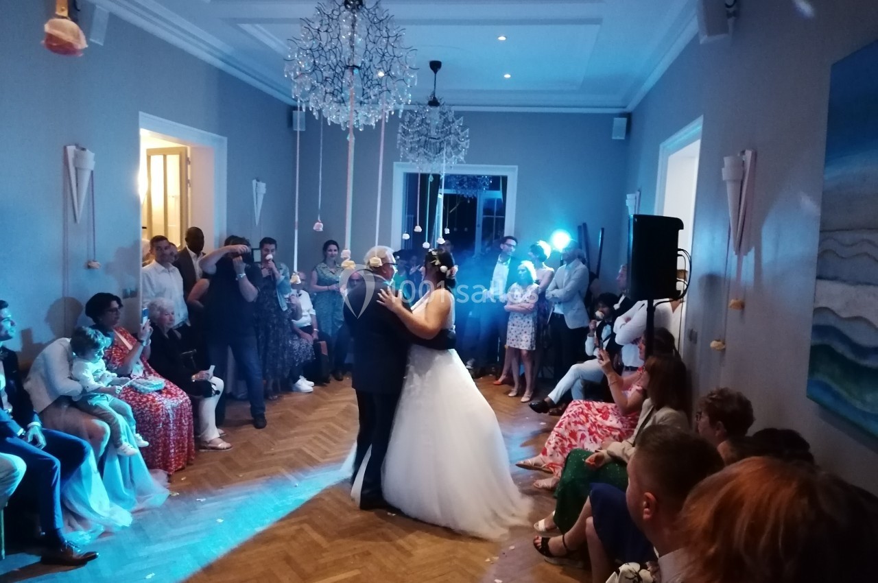 Un couple de mariés danse au centre d'une salle éclairée, entouré d'invités assis et debout.