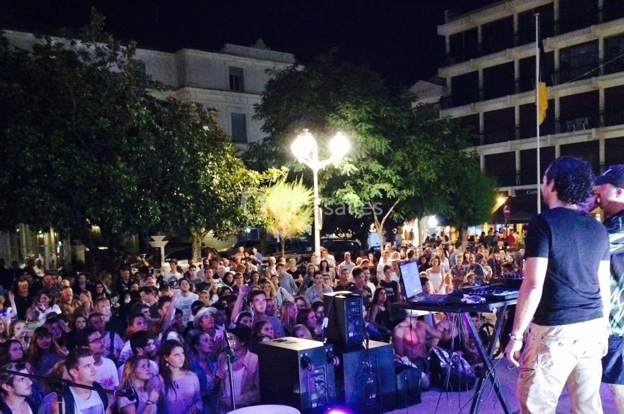 Une foule assiste à un concert en plein air de nuit, avec deux musiciens sur scène et des éclairages urbains.