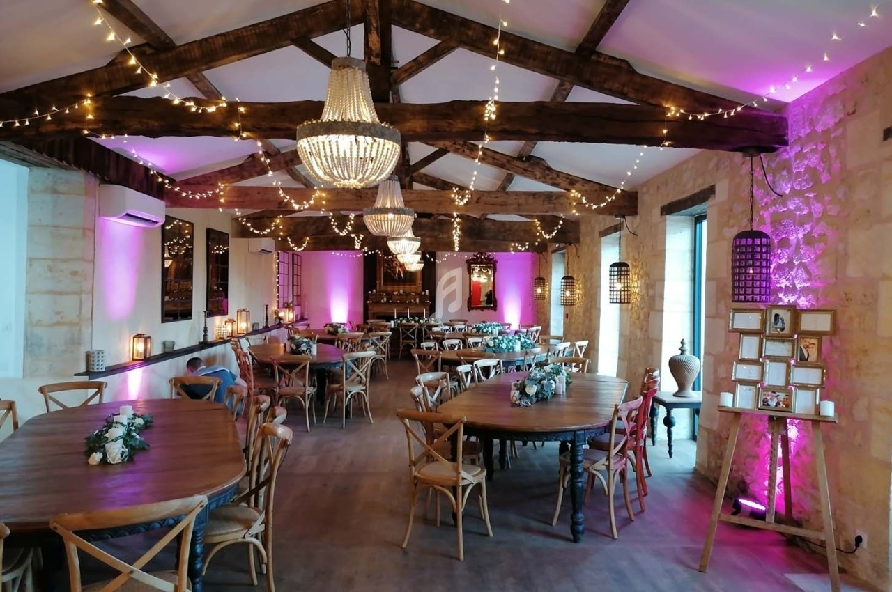 Salle de réception avec poutres apparentes, tables en bois décorées de fleurs et éclairage tamisé rose et blanc.