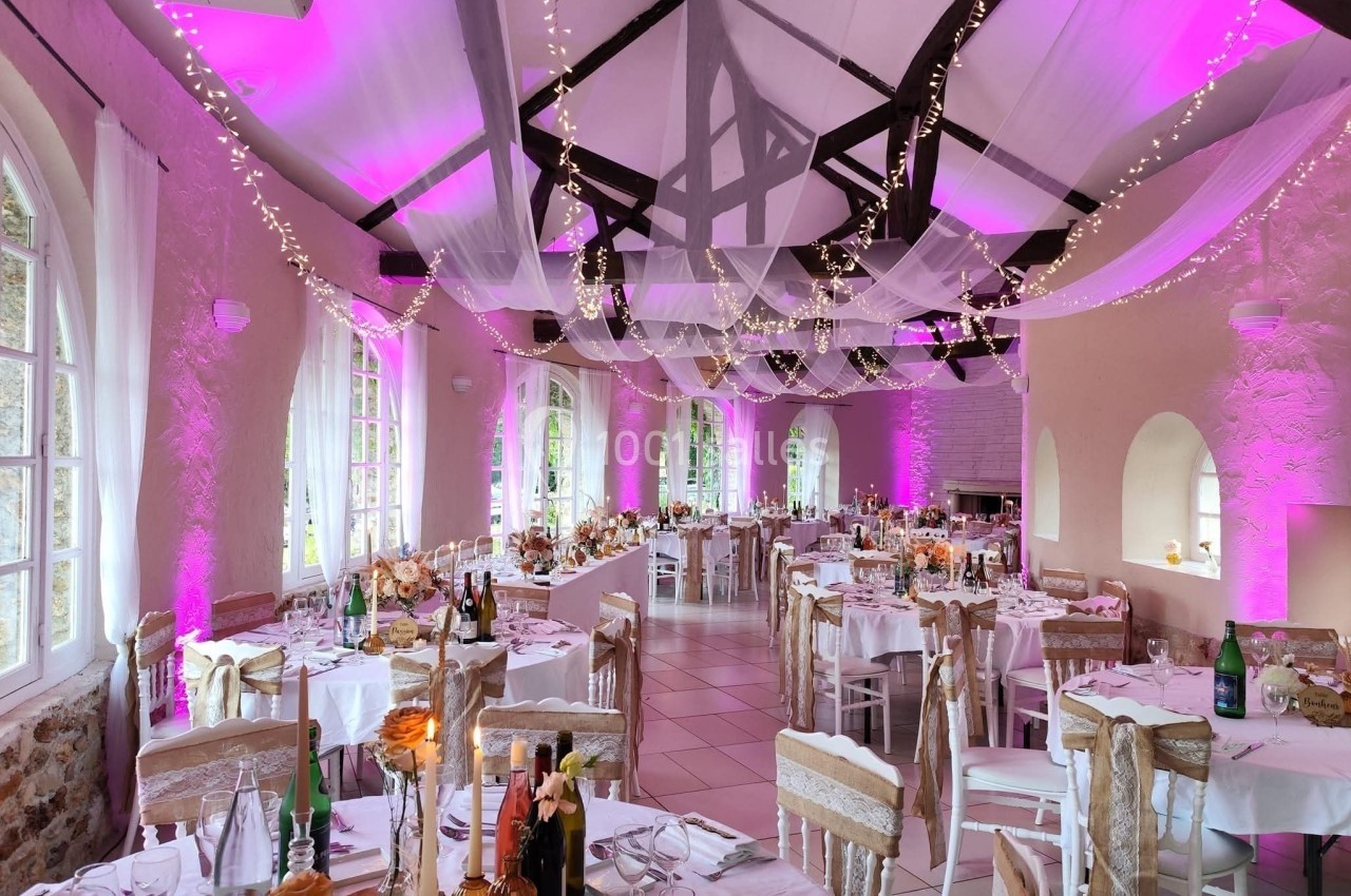 Salle de réception décorée avec des guirlandes lumineuses, des nappes blanches et une ambiance lumineuse rose.