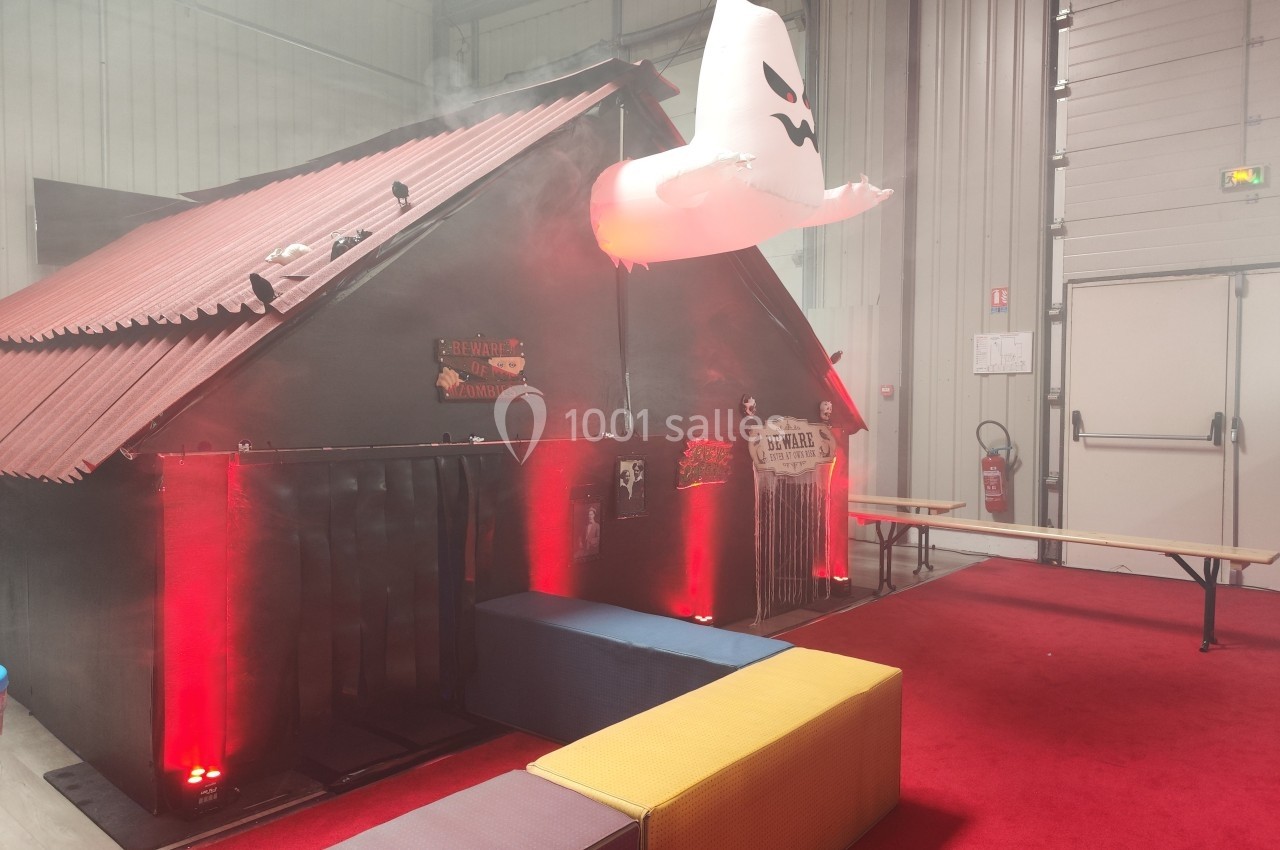 Cabane décorée pour Halloween avec éclairage rouge, fantôme suspendu et accessoires effrayants dans un espace intérieur.
