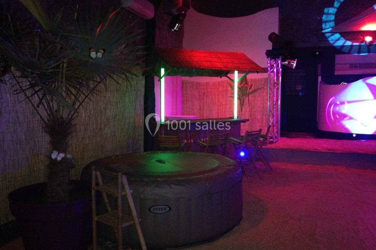 Un jacuzzi gonflable installé dans un espace intérieur décoré avec un bar en bois, des lumières colorées et du sable.