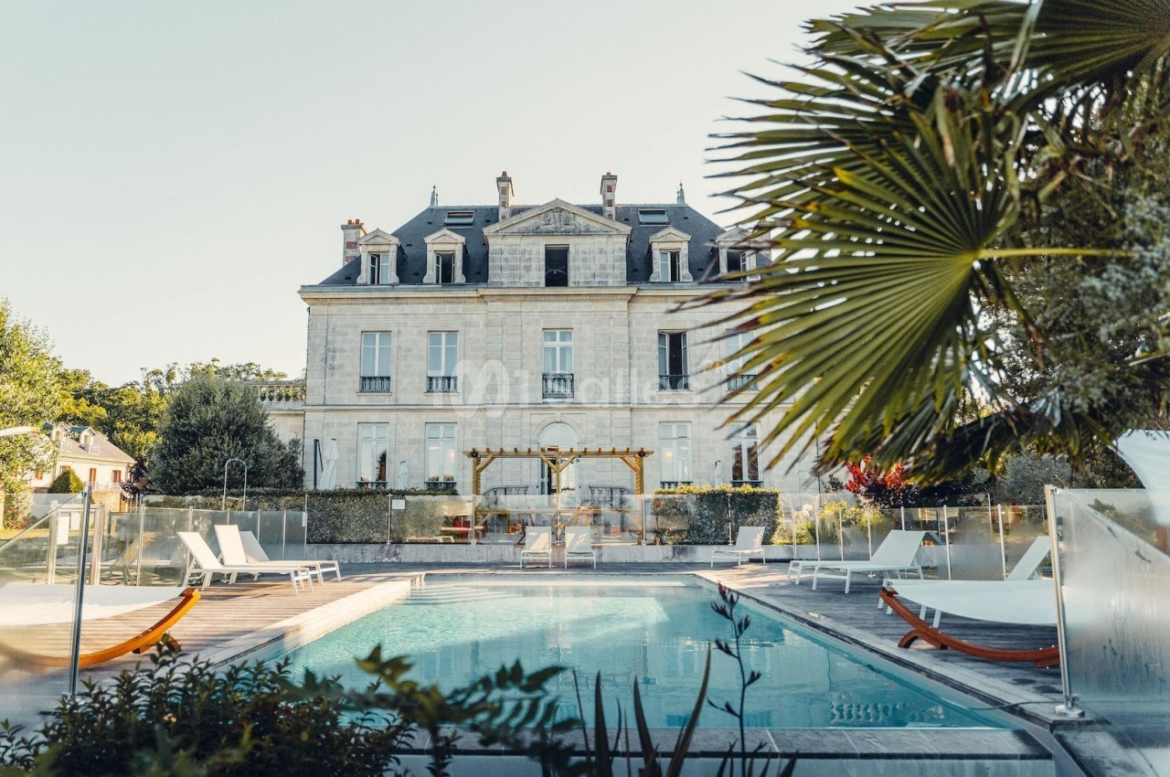 Façade d'un manoir en pierre avec piscine au premier plan, entouré de végétation et de chaises longues.