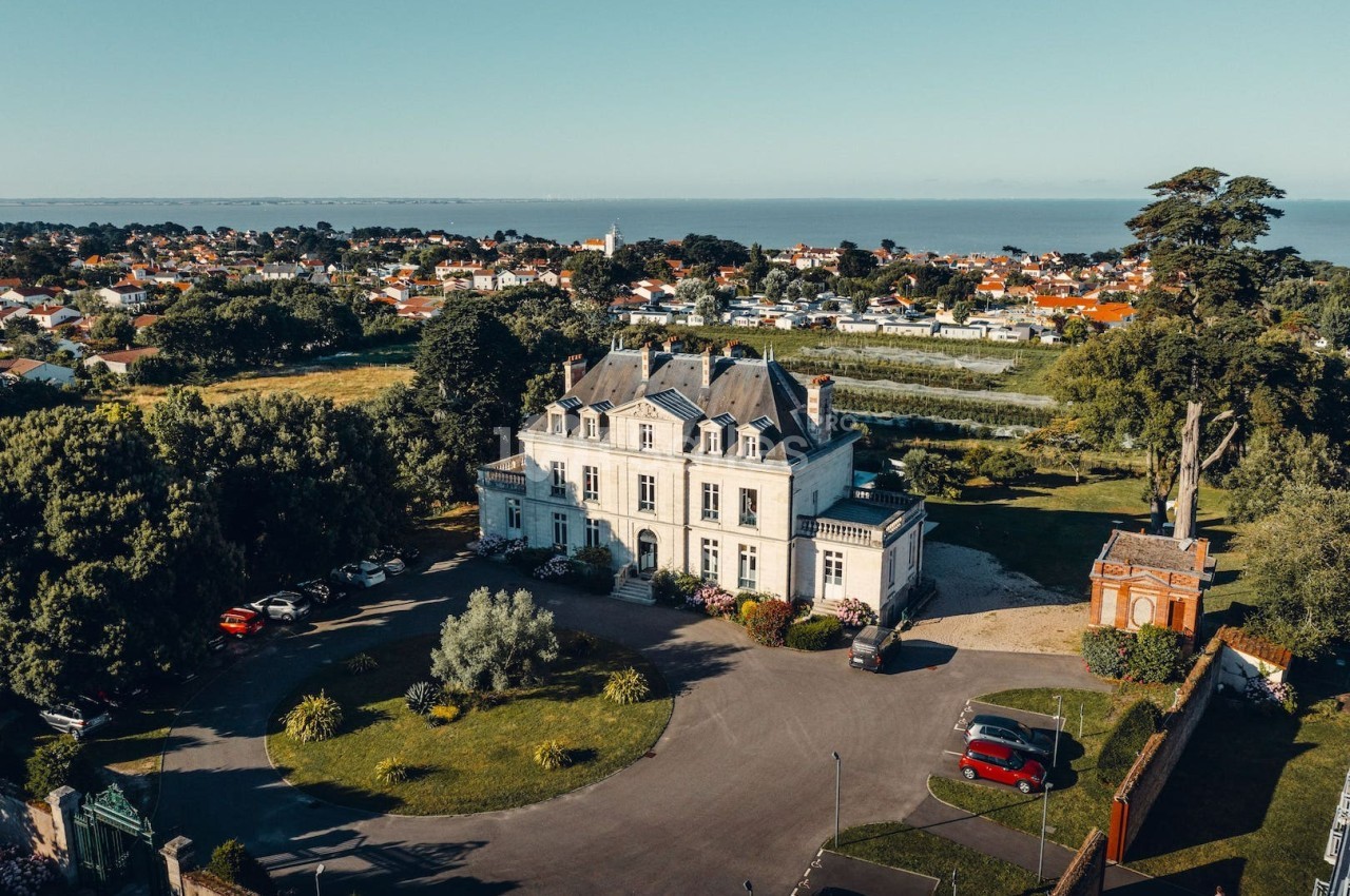 Vue aérienne d'un manoir entouré de jardins, avec des maisons et la mer en arrière-plan.