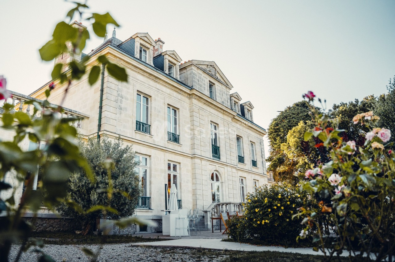 Façade d'un manoir en pierre entouré de jardins fleuris sous un ciel dégagé.
