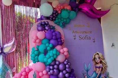 Arc décoratif en ballons roses, bleus et dorés avec papillons, installé dans une pièce au sol en bois.