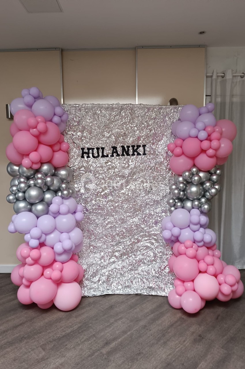 Mur décoratif avec fond argenté, encadré de ballons roses, violets et argentés, et le mot ’HULANKI’ inscrit au centre.