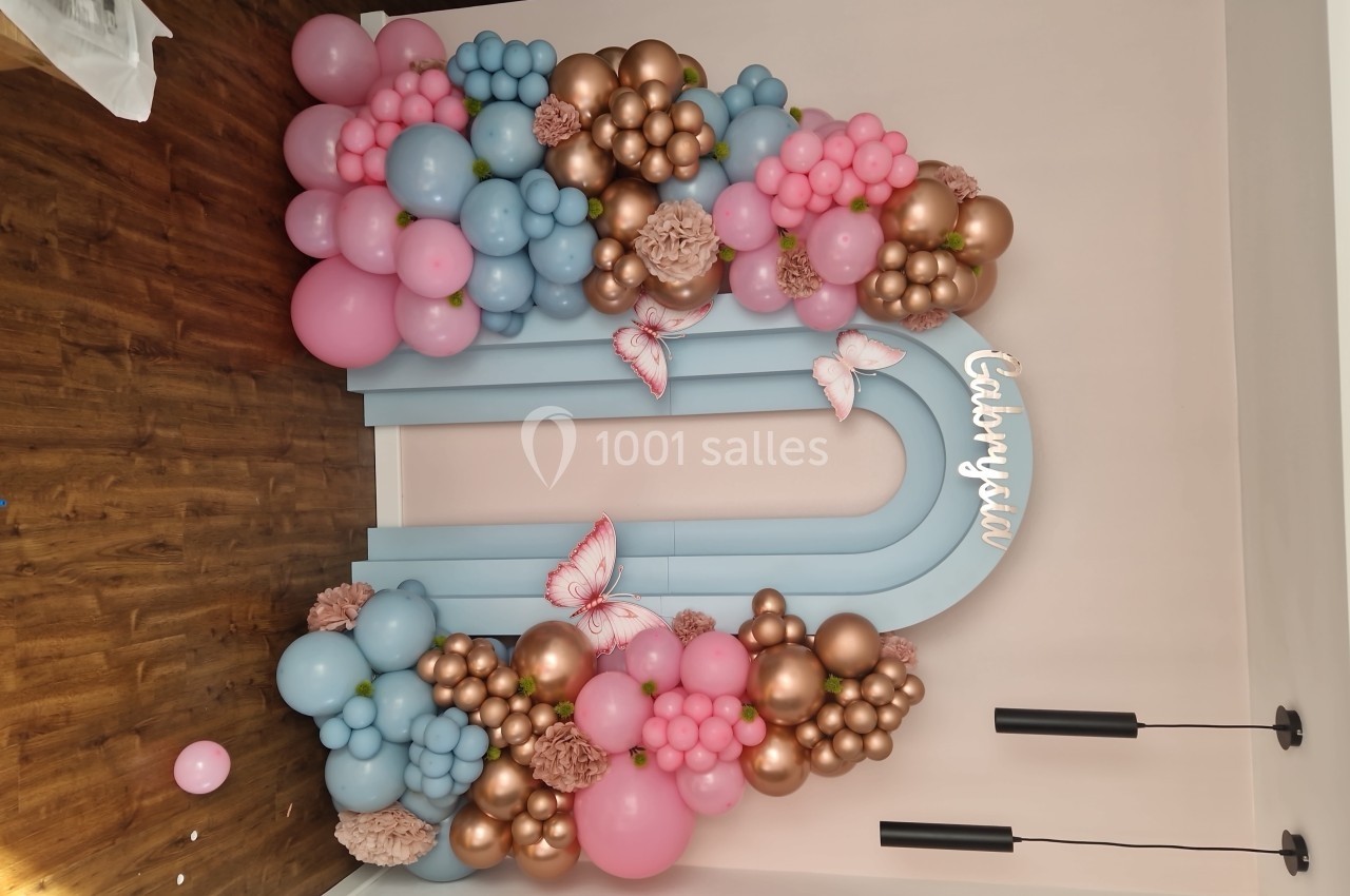 Arc décoratif en ballons roses, bleus et dorés avec papillons, installé dans une pièce au sol en bois.