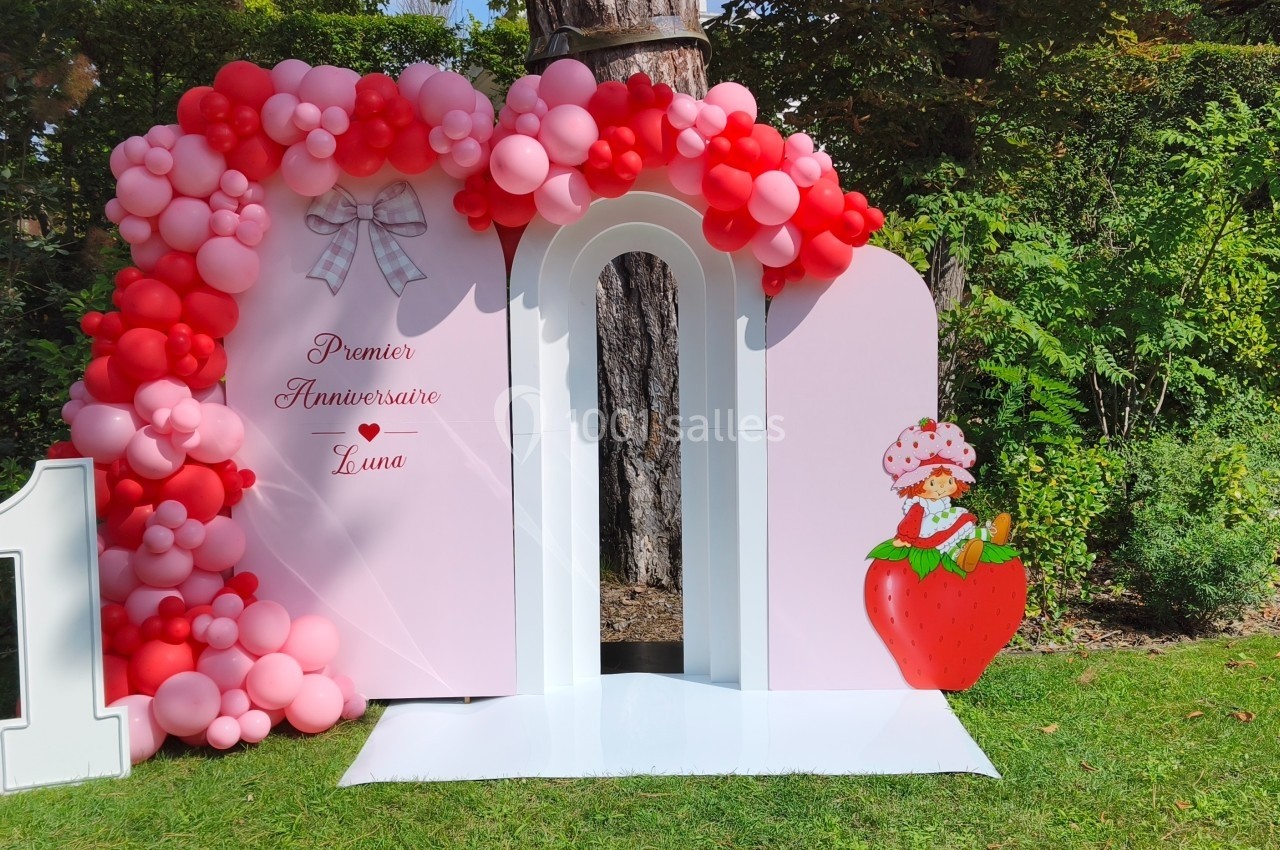 Décor d'anniversaire avec arche rose ornée de ballons rouges et roses, chiffre 1 et illustration de fraise.