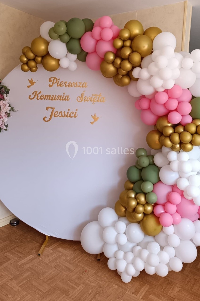 Décoration de communion avec arche de ballons colorés et panneau circulaire portant un message en polonais.