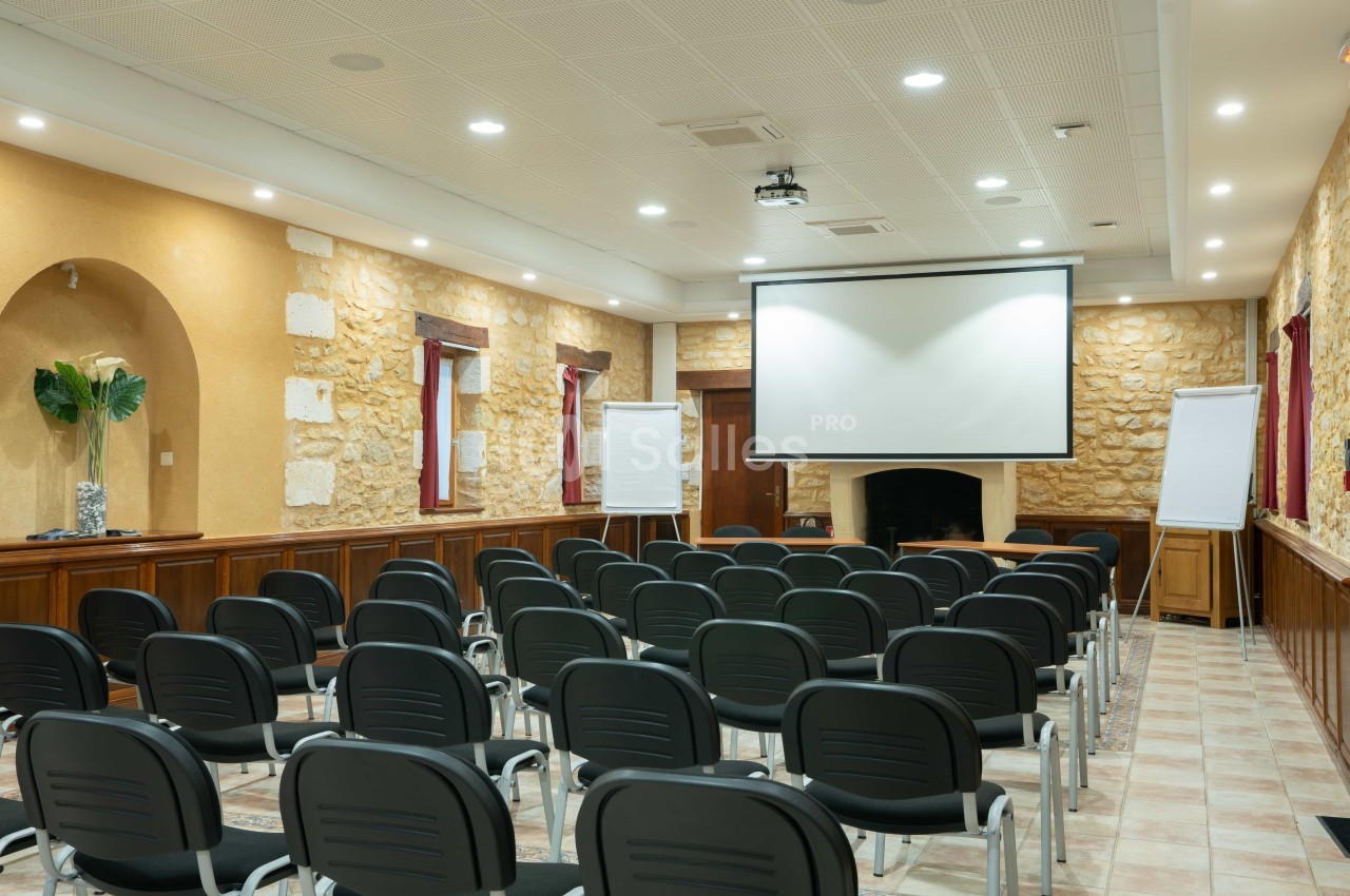 Salle de conférence avec des chaises alignées, un écran de projection, des tableaux blancs et des murs en pierre apparente.