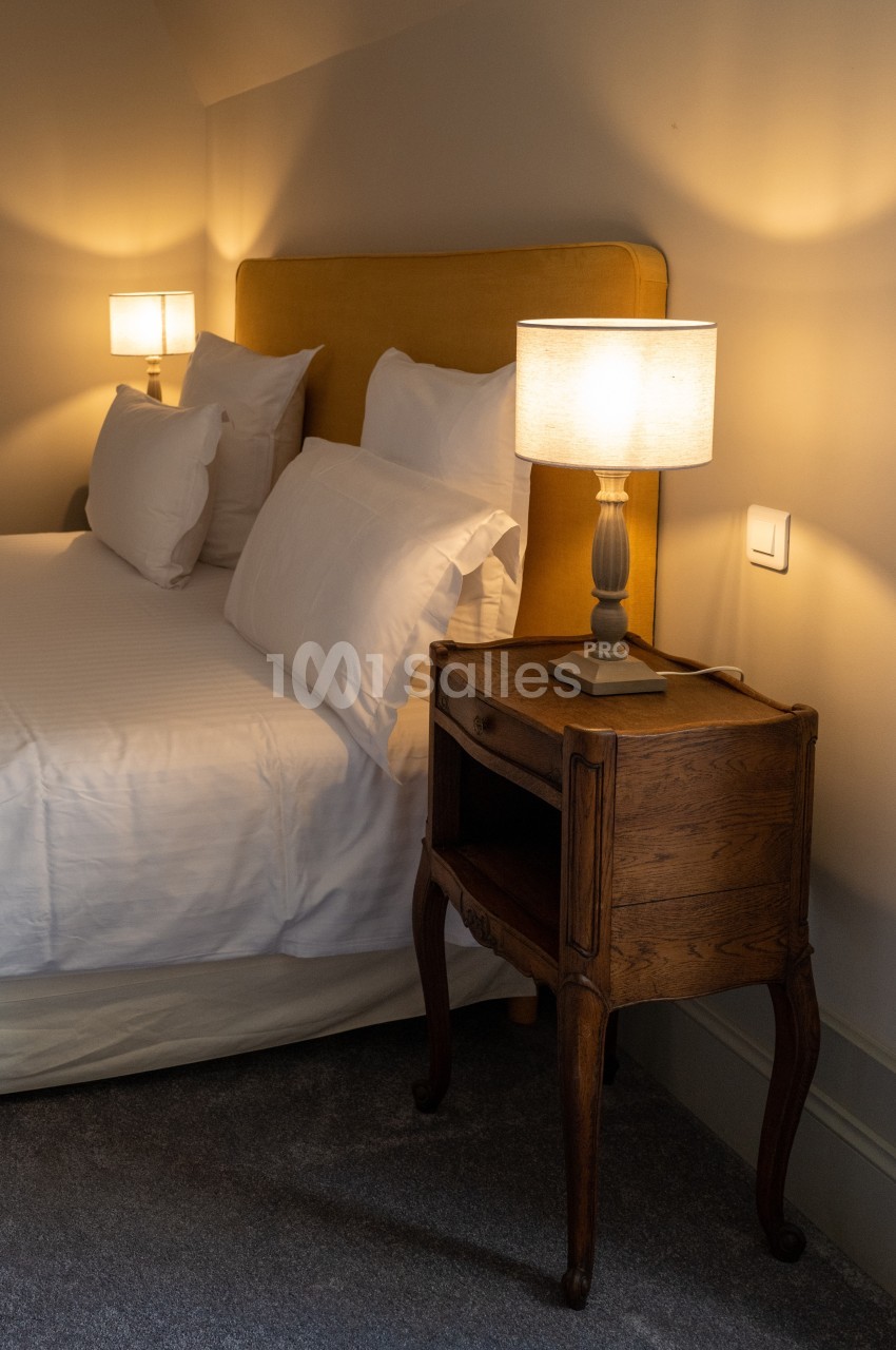 Table de chevet en bois avec lampe allumée, placée à côté d'un lit avec tête de lit jaune et draps blancs.