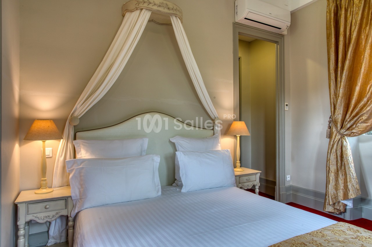 Lit double avec baldaquin, draps blancs, tables de chevet avec lampes allumées, rideaux dorés et mur beige.