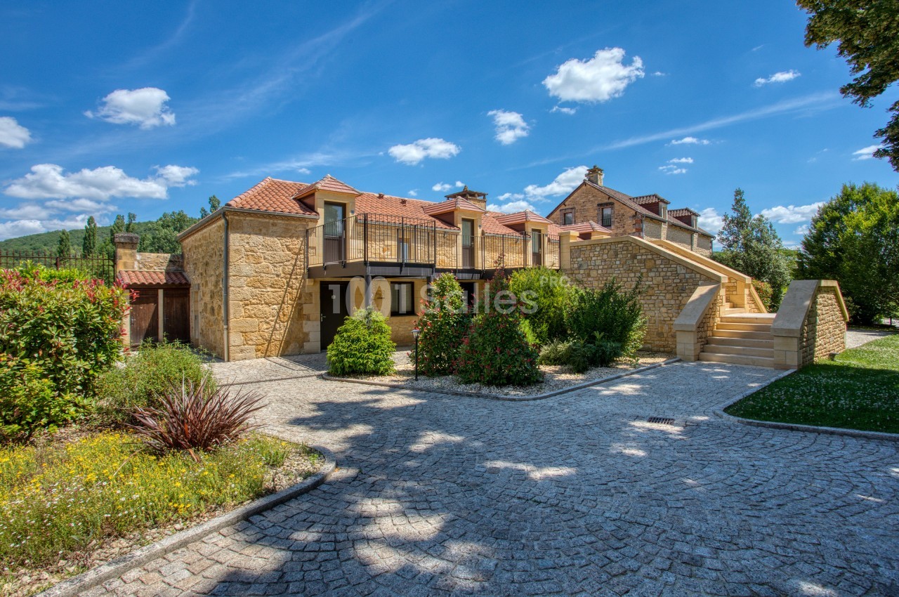 Maison en pierre avec toit en tuiles, entourée de végétation, sous un ciel bleu parsemé de nuages.