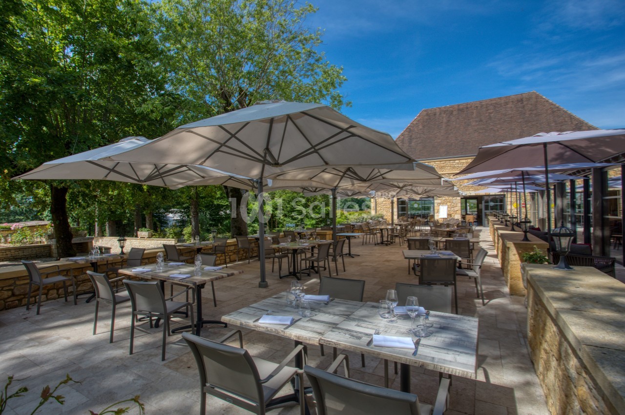 Terrasse d'un restaurant avec tables dressées, chaises et grands parasols, entourée de verdure et baignée de lumière…
