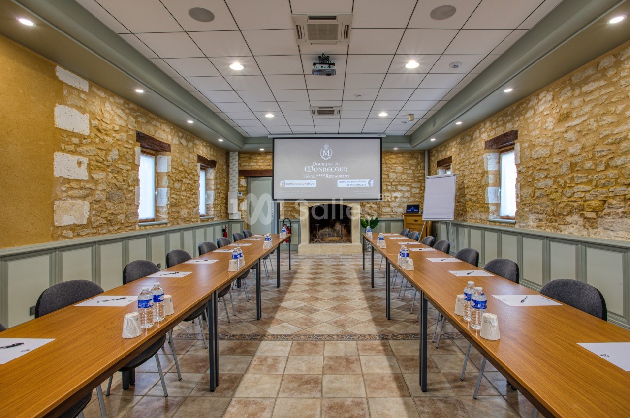 Salle de réunion avec murs en pierre, tables en U équipées de bouteilles d'eau, cahiers et stylos, écran de projection…