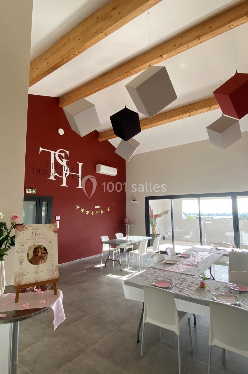 Salle lumineuse décorée pour un événement, avec tables dressées, cubes suspendus et mur rouge portant un logo.