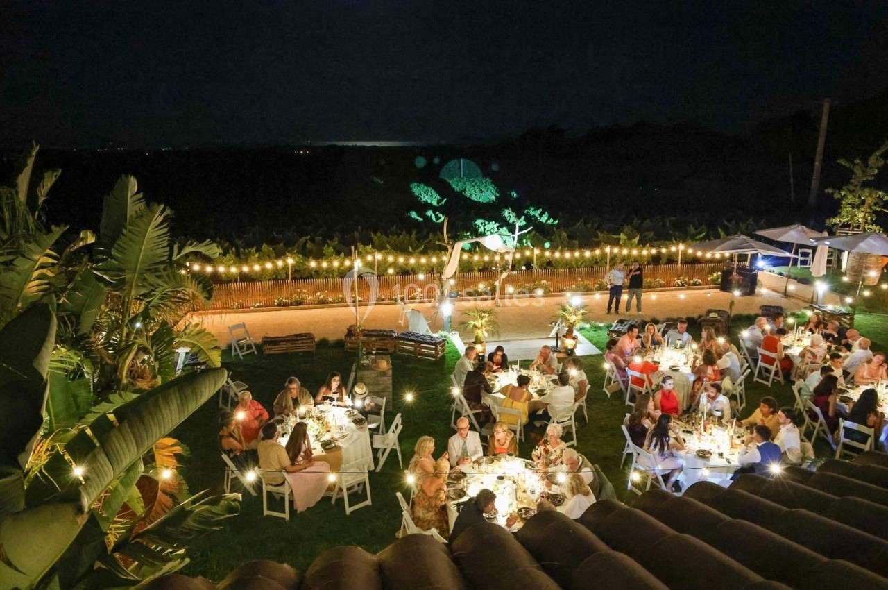 Vue nocturne d'un dîner en plein air avec des invités assis à des tables éclairées, entourés de végétation tropicale.
