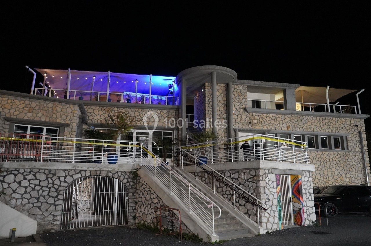 Bâtiment en pierre éclairé la nuit, avec terrasse illuminée en bleu et escalier central menant à l'entrée principale.
