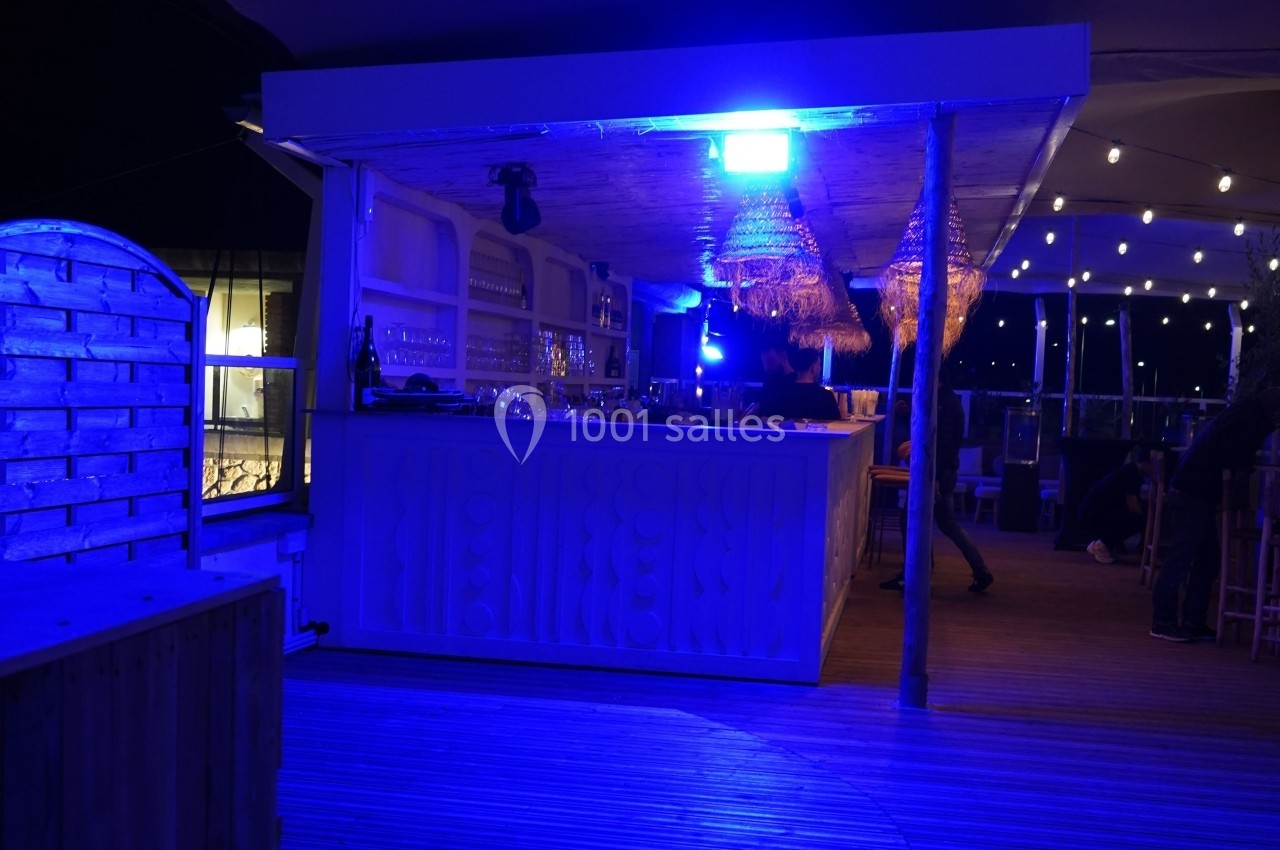 Bar extérieur éclairé par une lumière bleue, avec des suspensions en osier et une terrasse en bois.