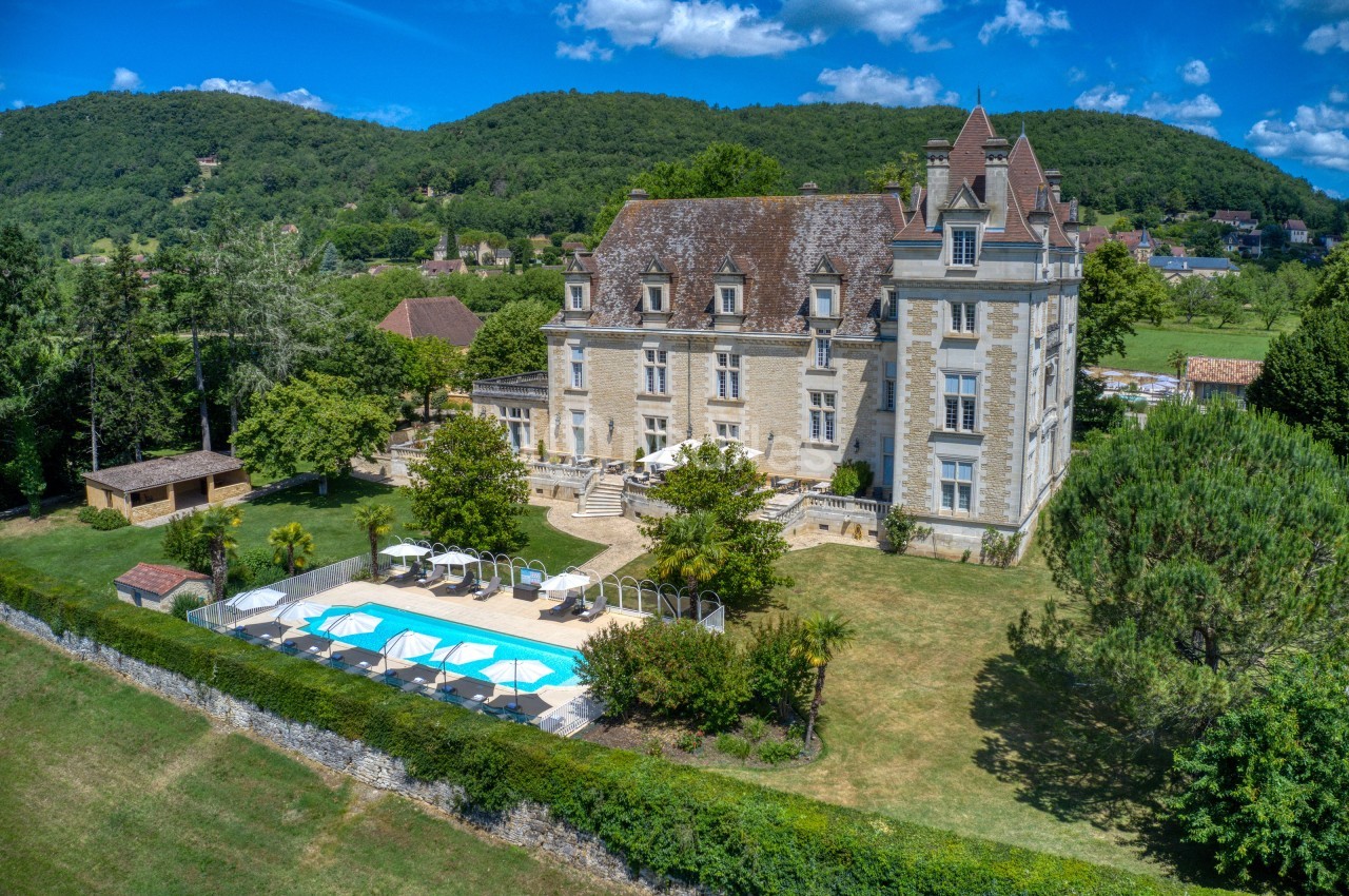 Vue aérienne d'un château en pierre entouré de verdure avec une piscine et des collines en arrière-plan.