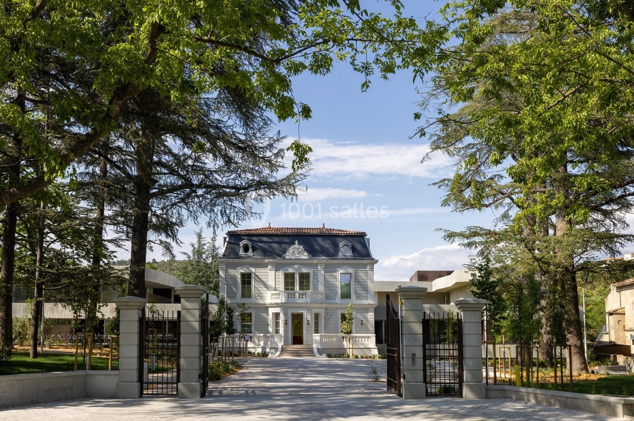 Façade d'une maison bourgeoise entourée d'arbres, avec une allée pavée et un portail ouvert au premier plan.