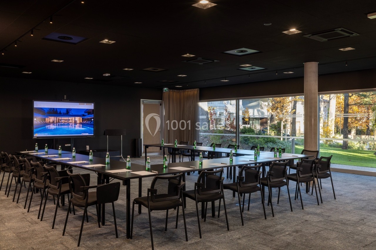 Salle de conférence moderne avec tables disposées en U, chaises noires, écran allumé et vue sur un jardin à travers de…