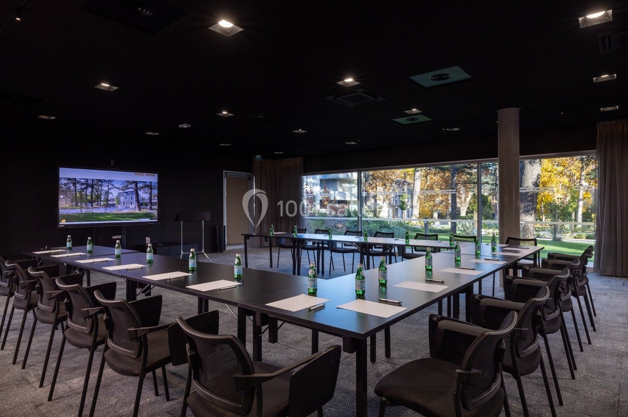 Salle de réunion moderne avec tables en U, chaises noires, bouteilles d'eau et vue sur un jardin à travers de grandes baies…