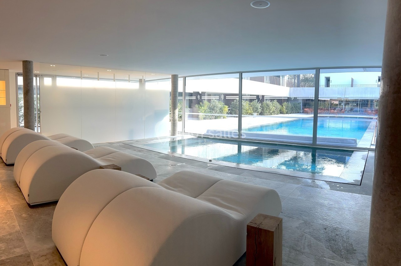Salle intérieure avec chaises longues blanches face à une piscine intérieure-extérieure donnant sur un espace extérieur…