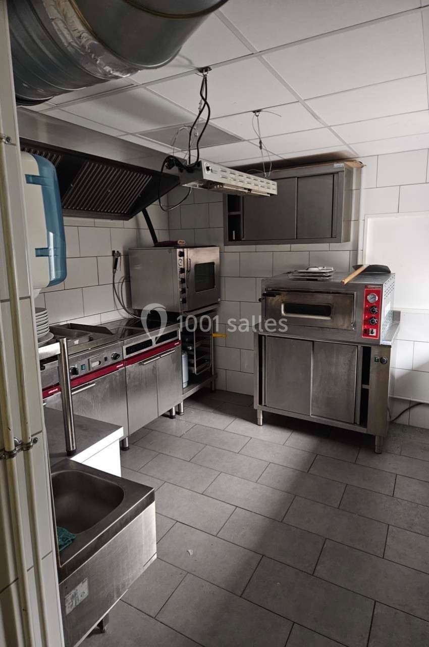 Cuisine professionnelle équipée avec four, hotte, évier et plan de travail en acier inoxydable, carrelage au sol et au mur.