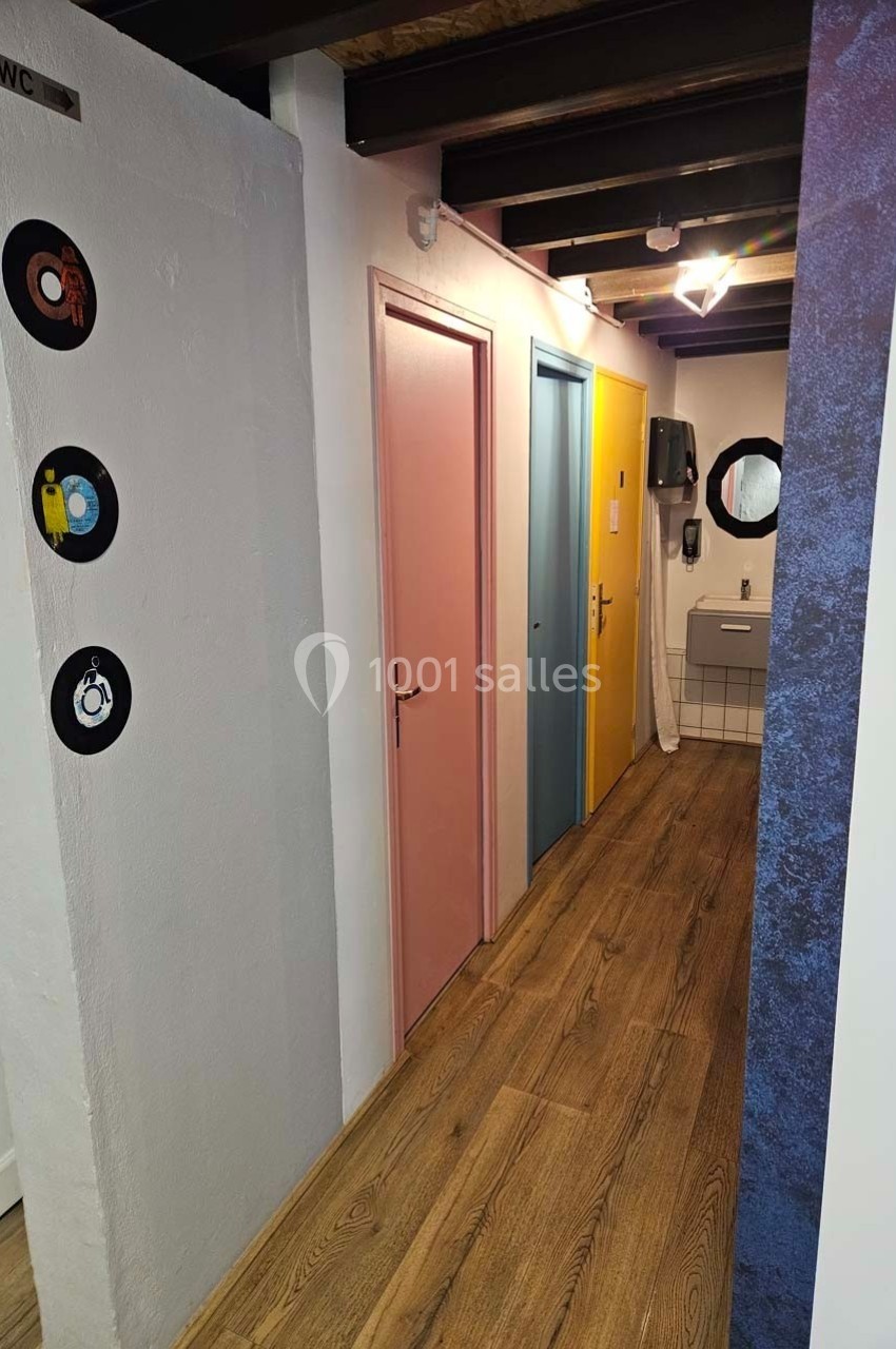 Couloir avec sol en bois, trois portes colorées (rose, bleu, jaune) et un lavabo avec miroir au fond.