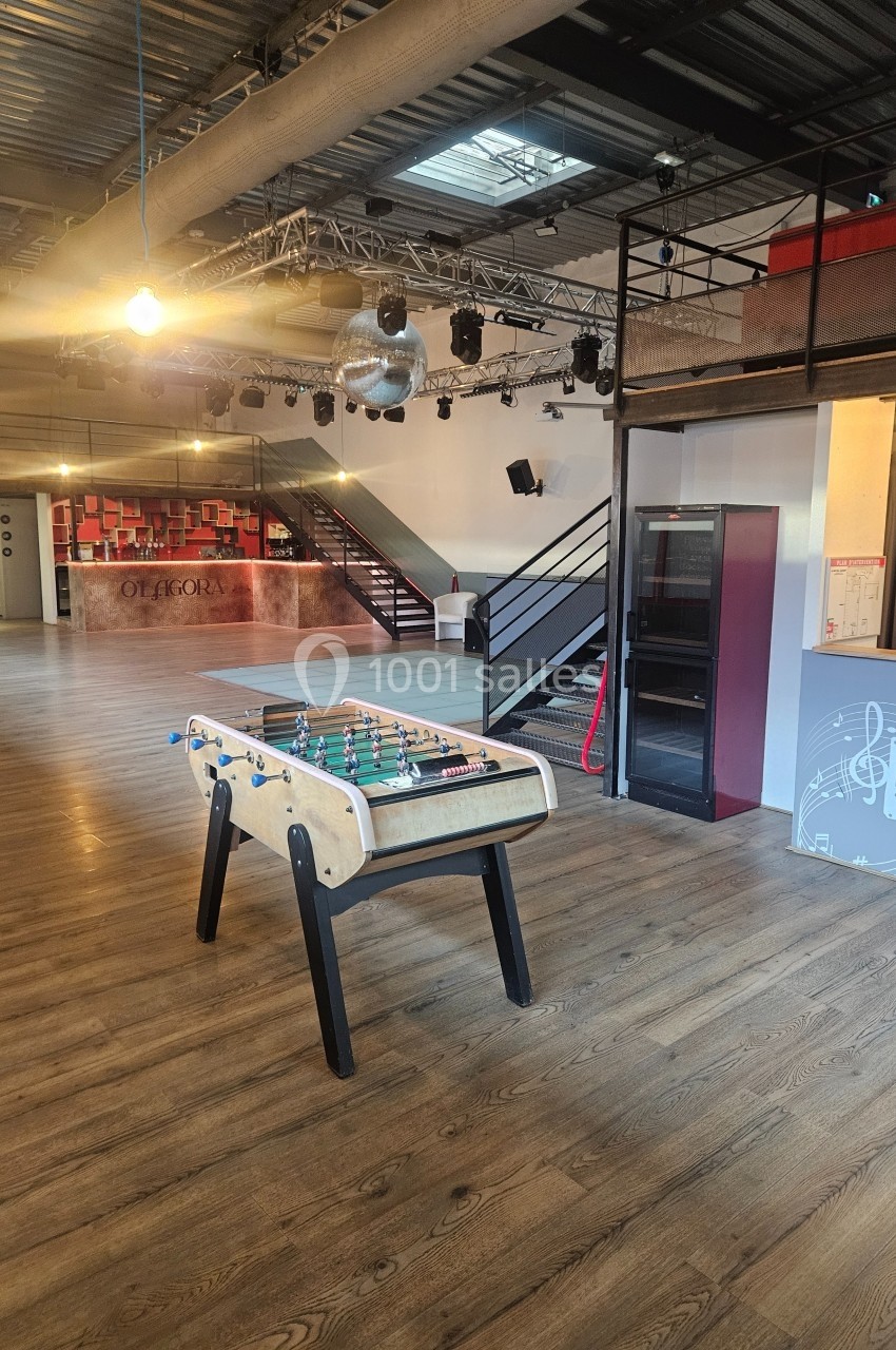 Salle spacieuse avec un baby-foot au premier plan, parquet en bois, escalier et éclairage industriel en arrière-plan.