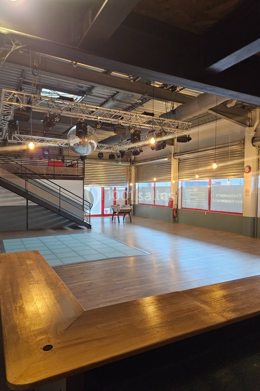 Salle vide avec parquet, piste de danse, escalier métallique et éclairage suspendu dans un espace industriel.