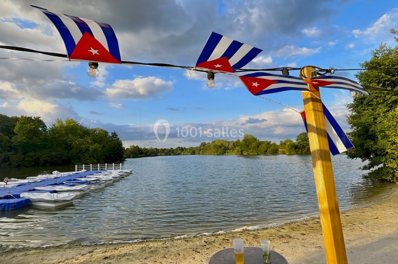 Drapeaux cubains accrochés à un poteau en bois près d'un lac avec des pédalos et une plage bordée d'arbres.