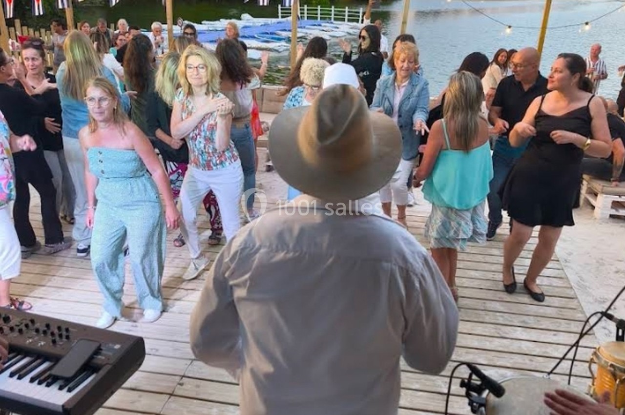 Groupe de personnes dansant sur une terrasse en bois près d'un lac, avec des musiciens en arrière-plan.