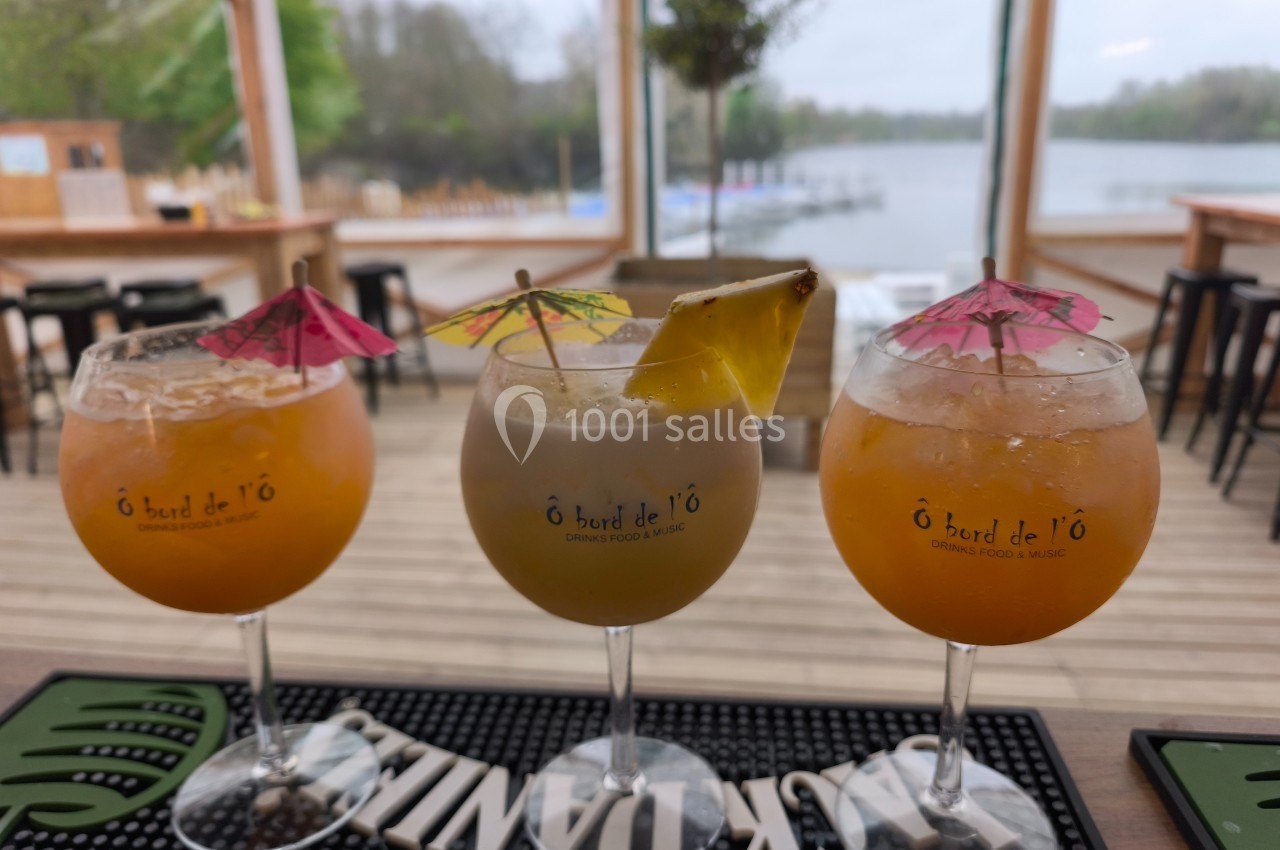 Trois cocktails colorés décorés de petits parasols et fruits, posés sur un bar avec vue sur un lac en arrière-plan.