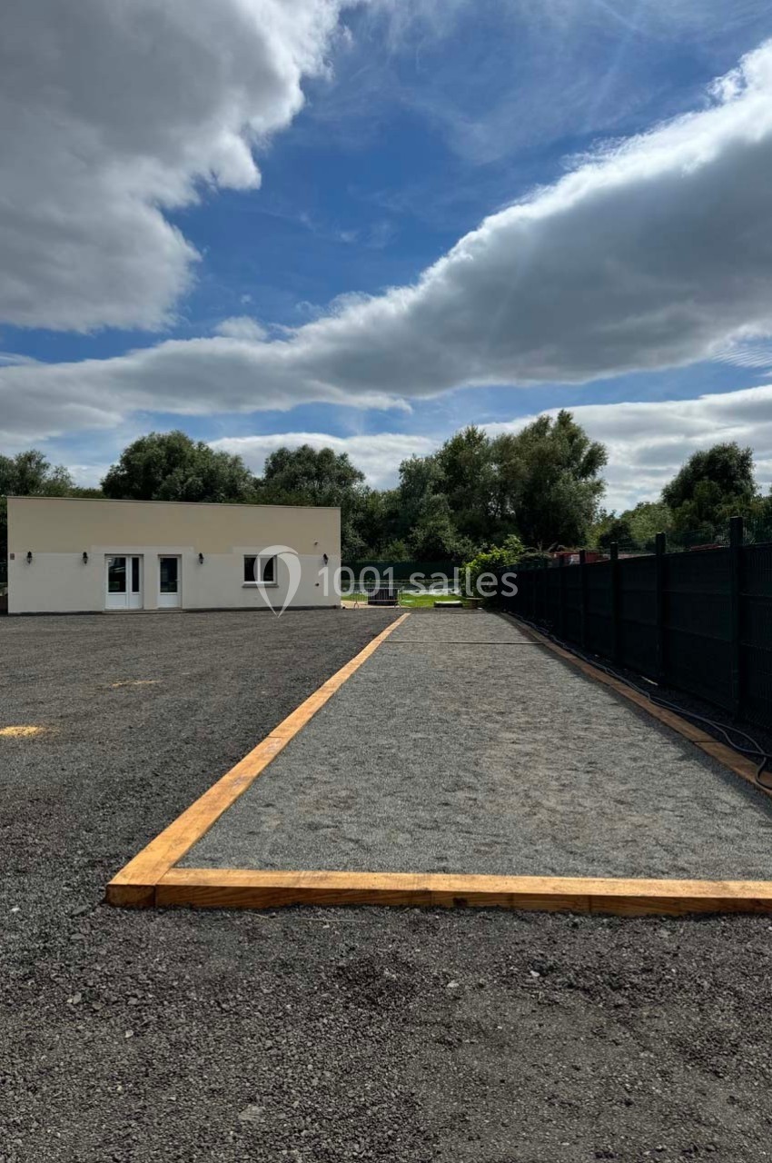 Terrain de pétanque en gravier bordé de bois, situé près d'un bâtiment blanc et entouré de verdure.