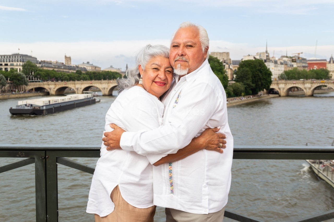 Un couple âgé s'enlace sur un pont avec une vue sur la Seine et des bâtiments parisiens en arrière-plan.