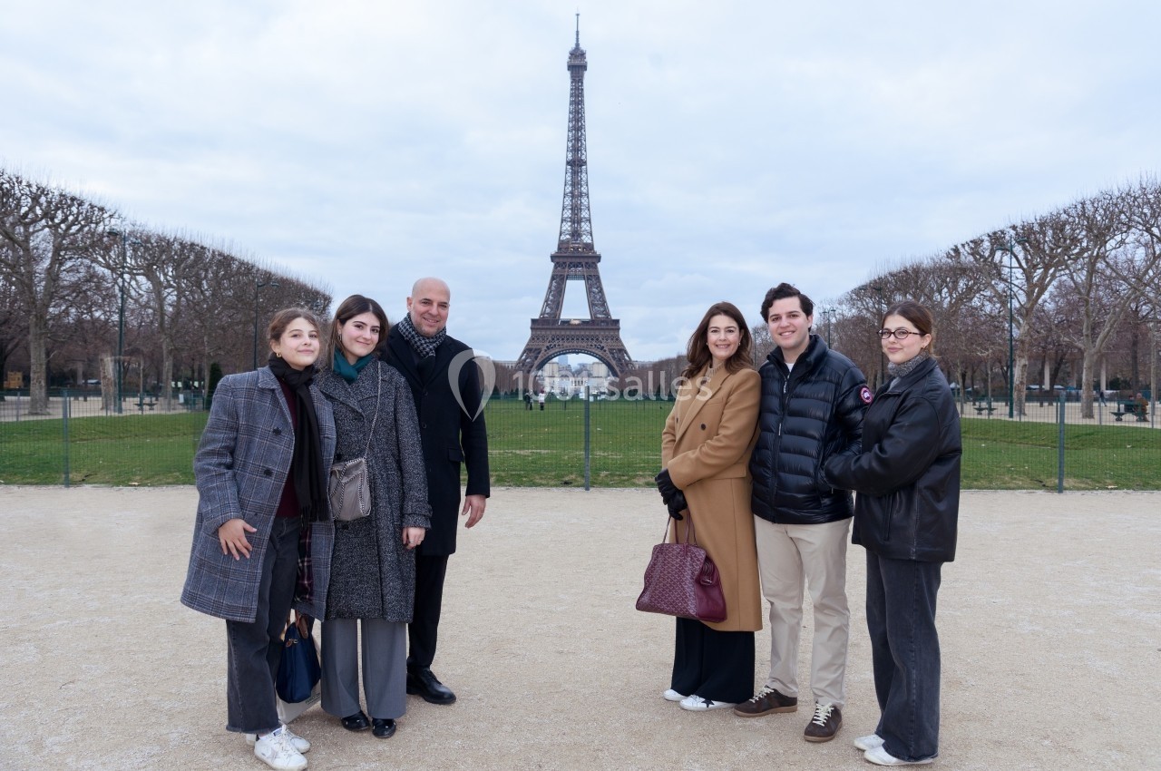 Un groupe de personnes pose devant la tour Eiffel par temps nuageux, dans un espace dégagé bordé d'arbres.