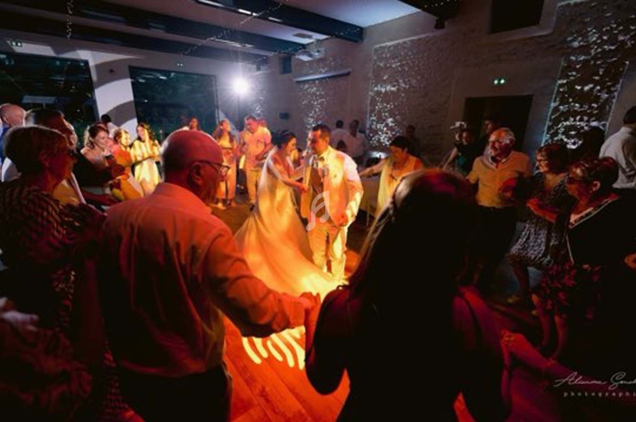 Des invités dansent en cercle autour des mariés dans une salle éclairée par des lumières tamisées et colorées.