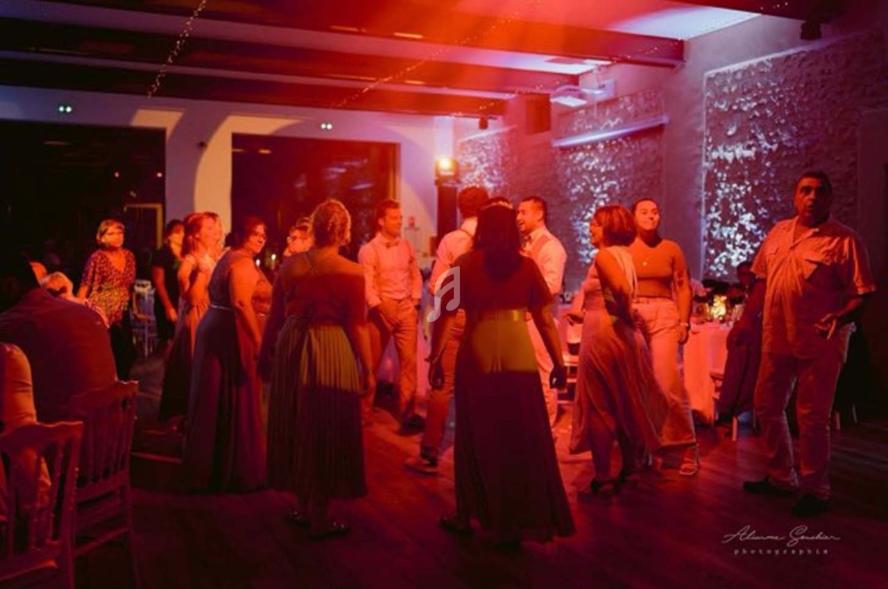 Des personnes dansent dans une salle éclairée par des lumières rouges et blanches, avec des tables en arrière-plan.