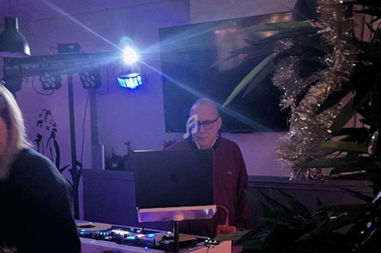 Un homme se tient derrière une table de mixage avec un ordinateur, dans une pièce faiblement éclairée décorée de plantes.