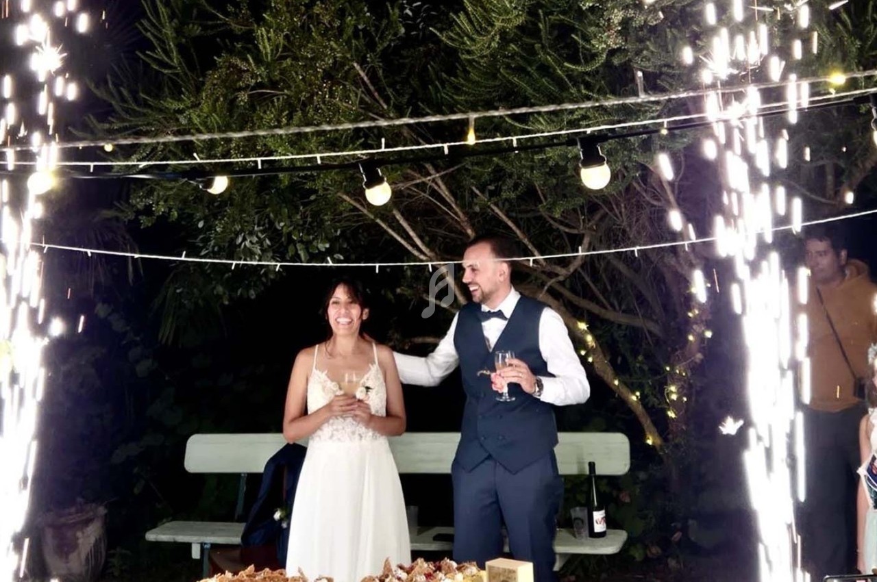 Un couple en tenue de mariage sourit sous des guirlandes lumineuses et des étincelles, devant une table décorée.