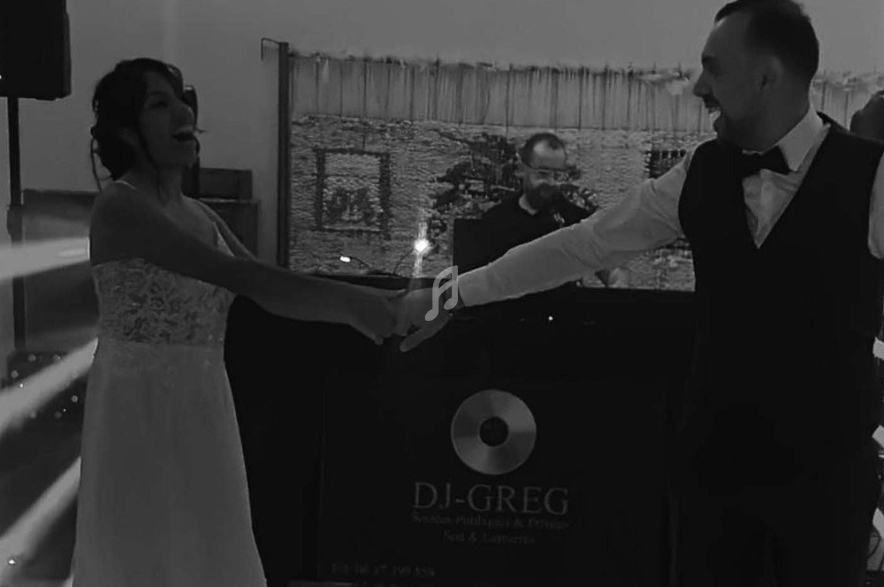 Un couple danse main dans la main devant un DJ lors d'une réception, éclairée par des jeux de lumière.