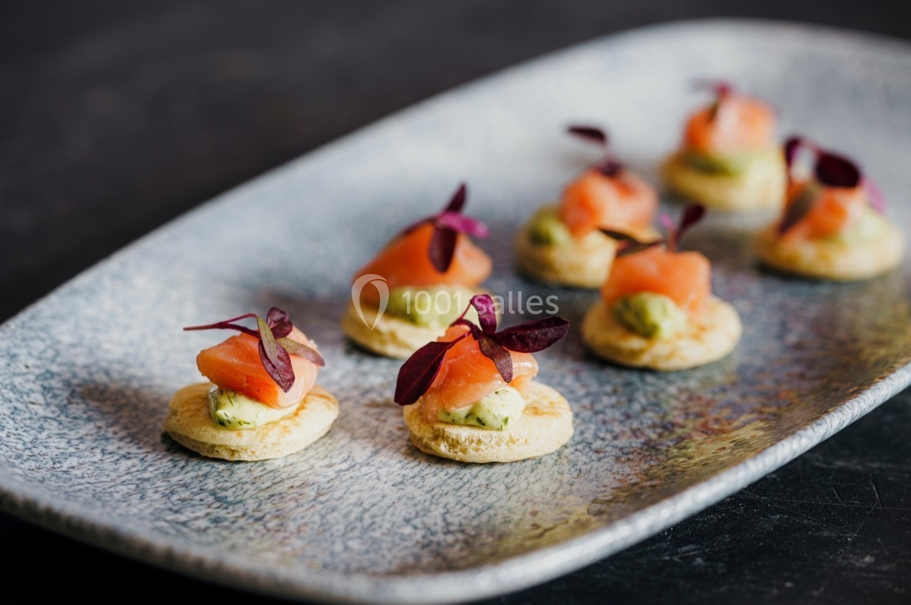 Assortiment de canapés au saumon fumé, crème d'avocat et feuilles décoratives sur un plateau gris.