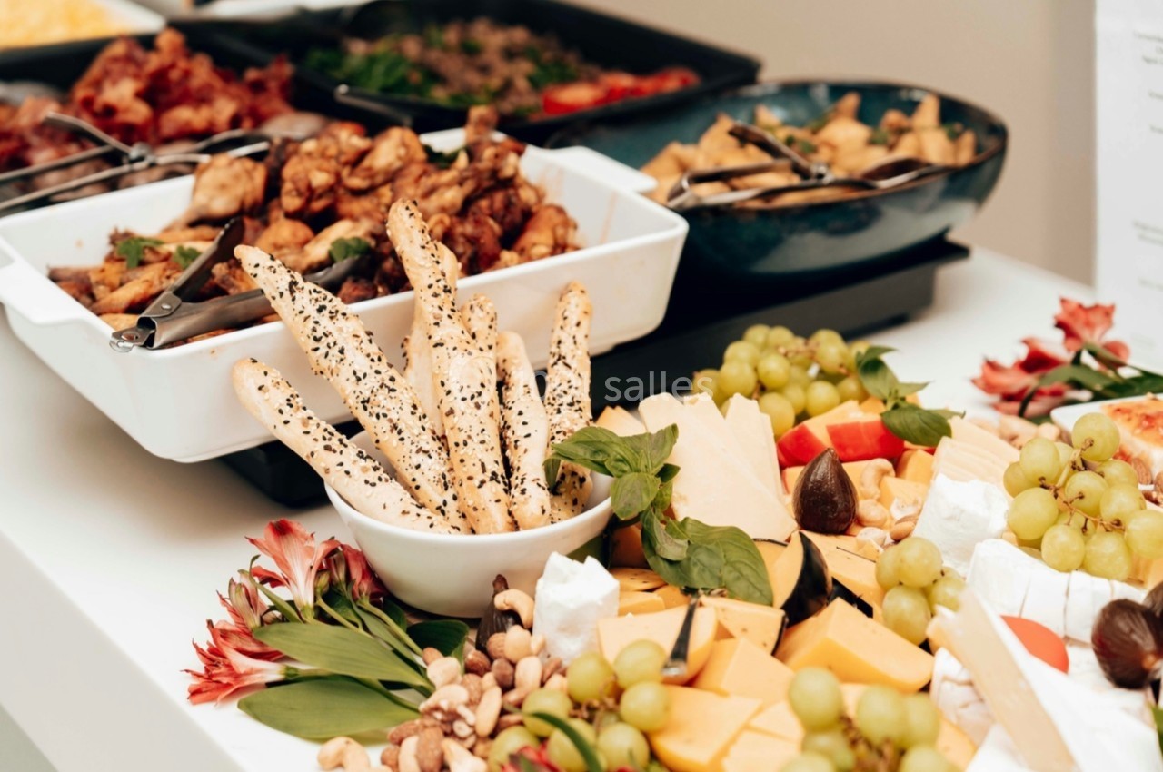 Plateau de buffet avec fromages, fruits, gressins aux graines, noix et plats variés disposés sur une table blanche.