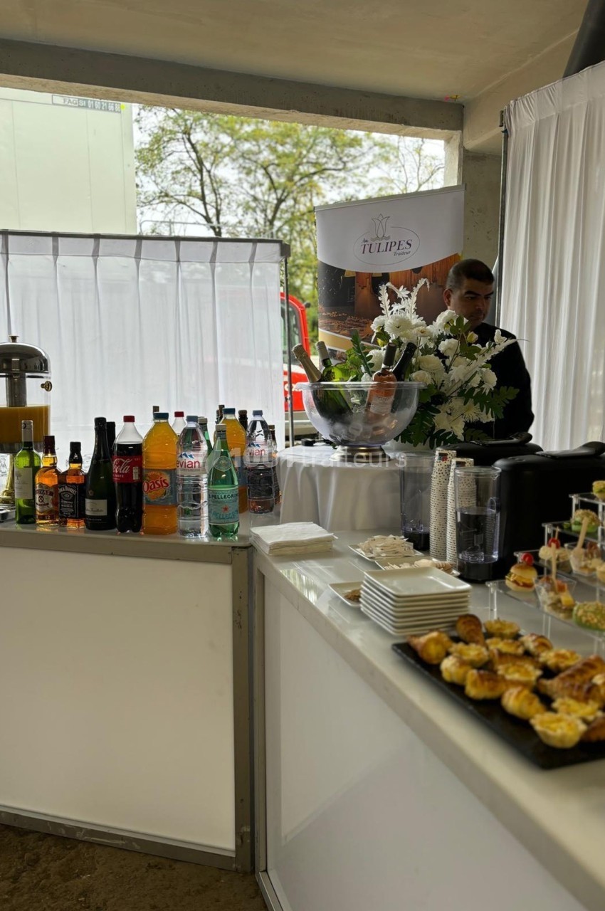 Buffet avec boissons variées, viennoiseries et fleurs blanches, installé dans un espace lumineux avec rideaux blancs.