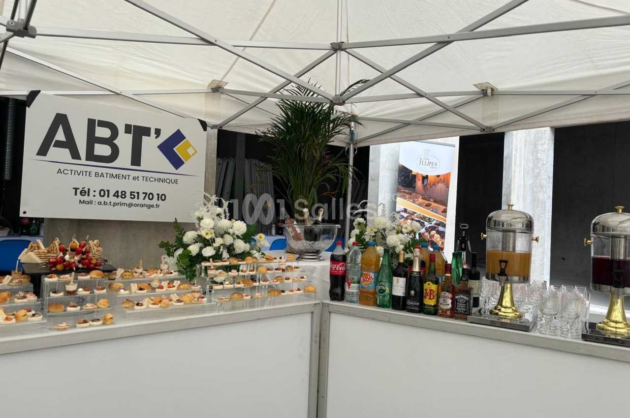Buffet sous une tente avec amuse-bouches, boissons variées et décoration florale, devant un panneau d'entreprise.